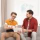 Two young men sit on a grey sofa looking at a touchscreen tablet device.